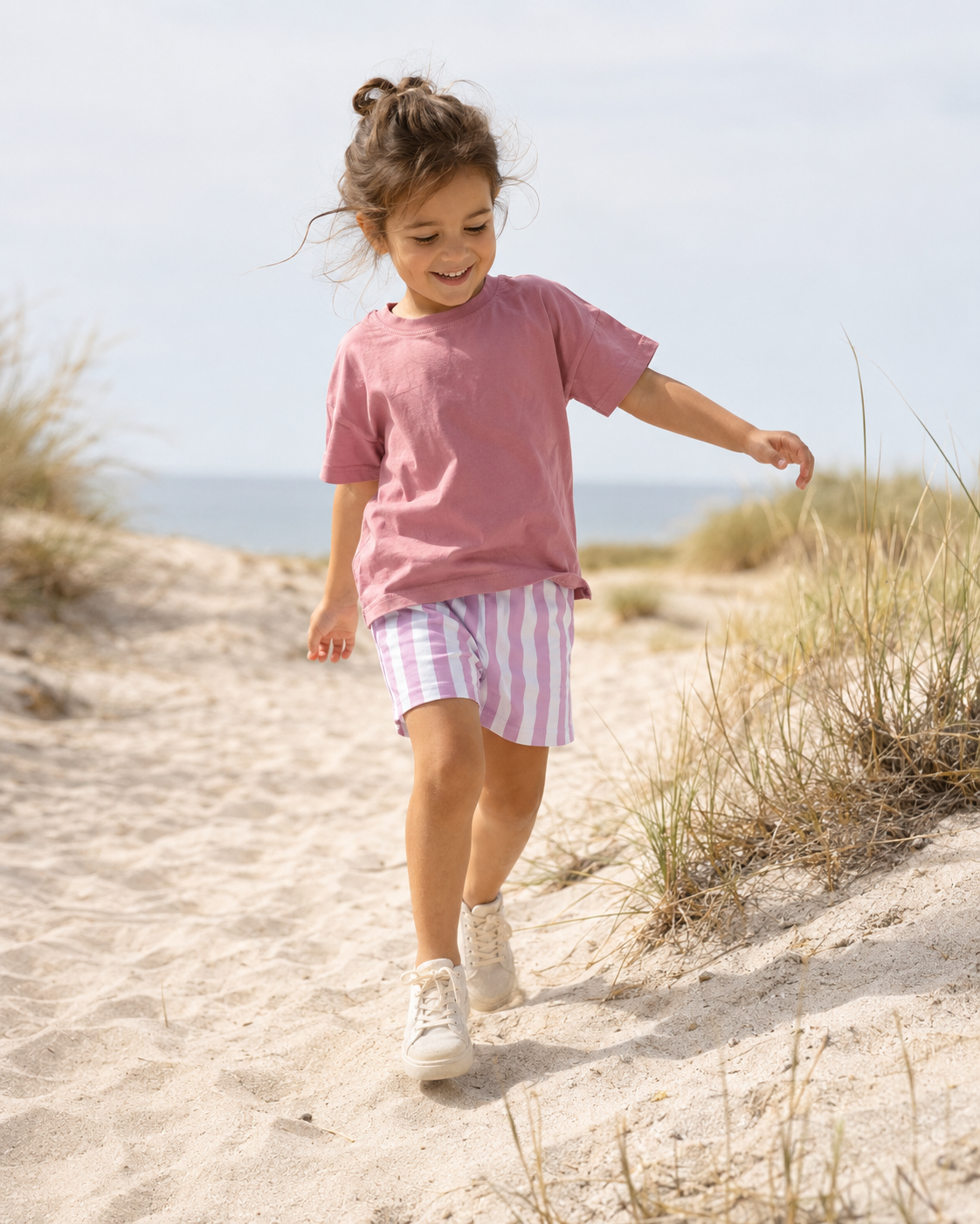 A young girl walking up sandy dunes near the beach, wearing a dusty pink t-shirt, pink and white striped shorts, and white sneakers.