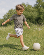 A young boy wearing a dark gray t-shirt, white and gray striped shorts, and white sneakers is running on a green grassy field while kicking a white soccer ball.