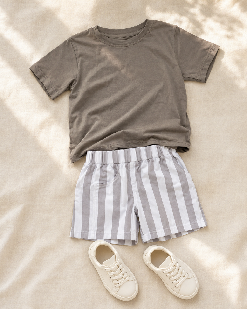 A styled flat-lay outfit featuring a short-sleeve crewneck t-shirt in taupe, striped shorts in gray and white, and a pair of cream-colored canvas sneakers, arranged on a beige fabric background with soft leaf shadows.