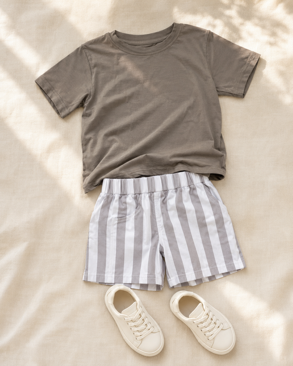 A styled flat-lay outfit featuring a short-sleeve crewneck t-shirt in taupe, striped shorts in gray and white, and a pair of cream-colored canvas sneakers, arranged on a beige fabric background with soft leaf shadows.